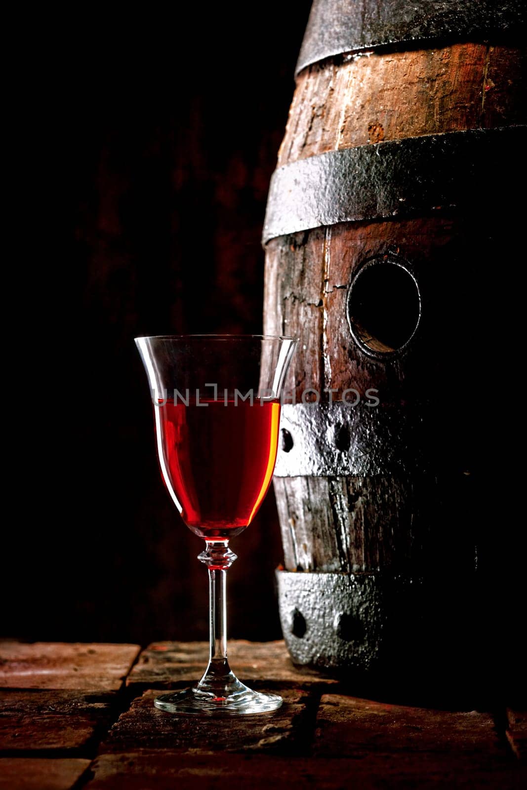 A glass of red wine next to an old oak cask with its stopper out.