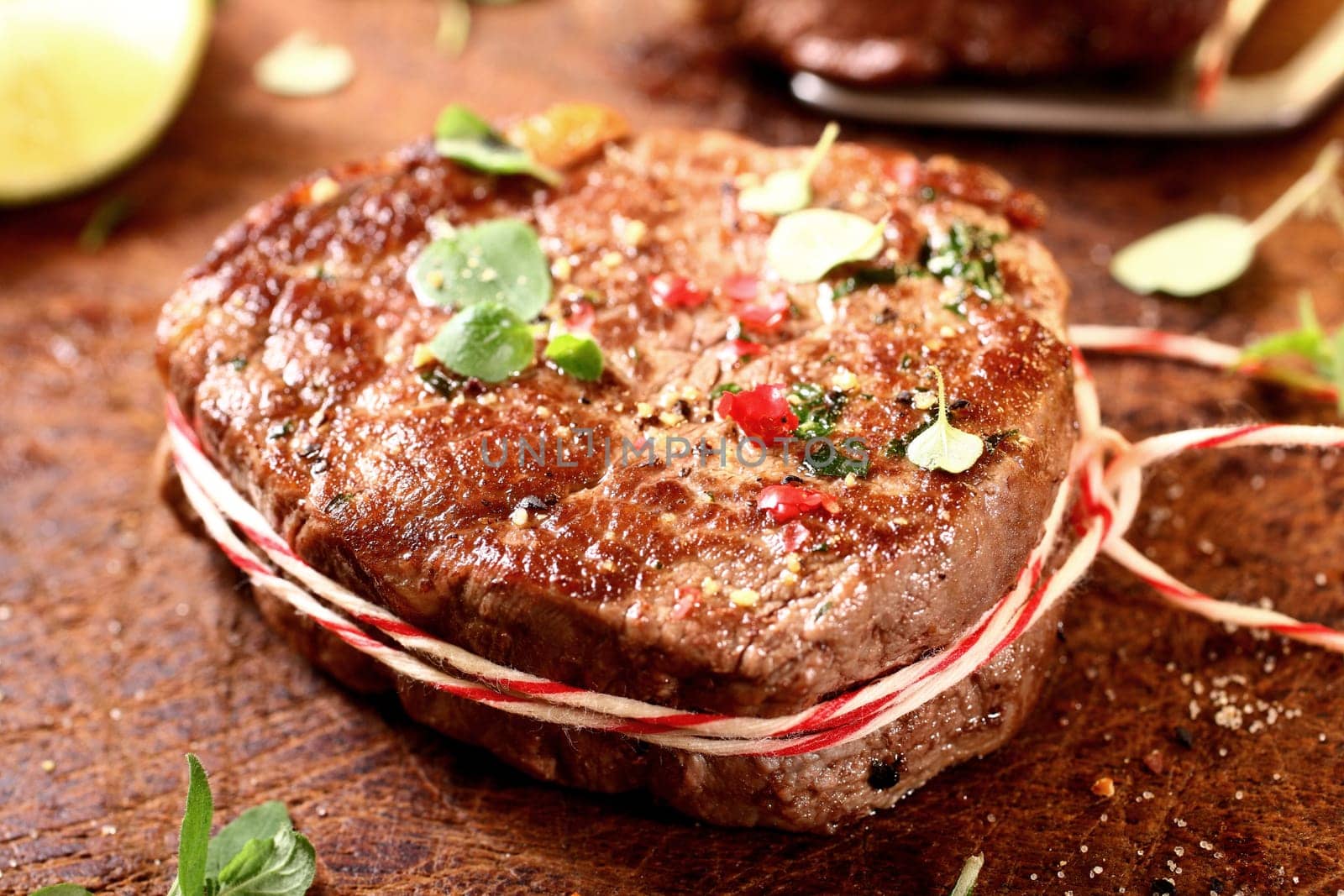 Thick juicy grilled lean beef steak medallions tied with colourful string and seasoned with chopped fresh herbs and spices on an old wooden board