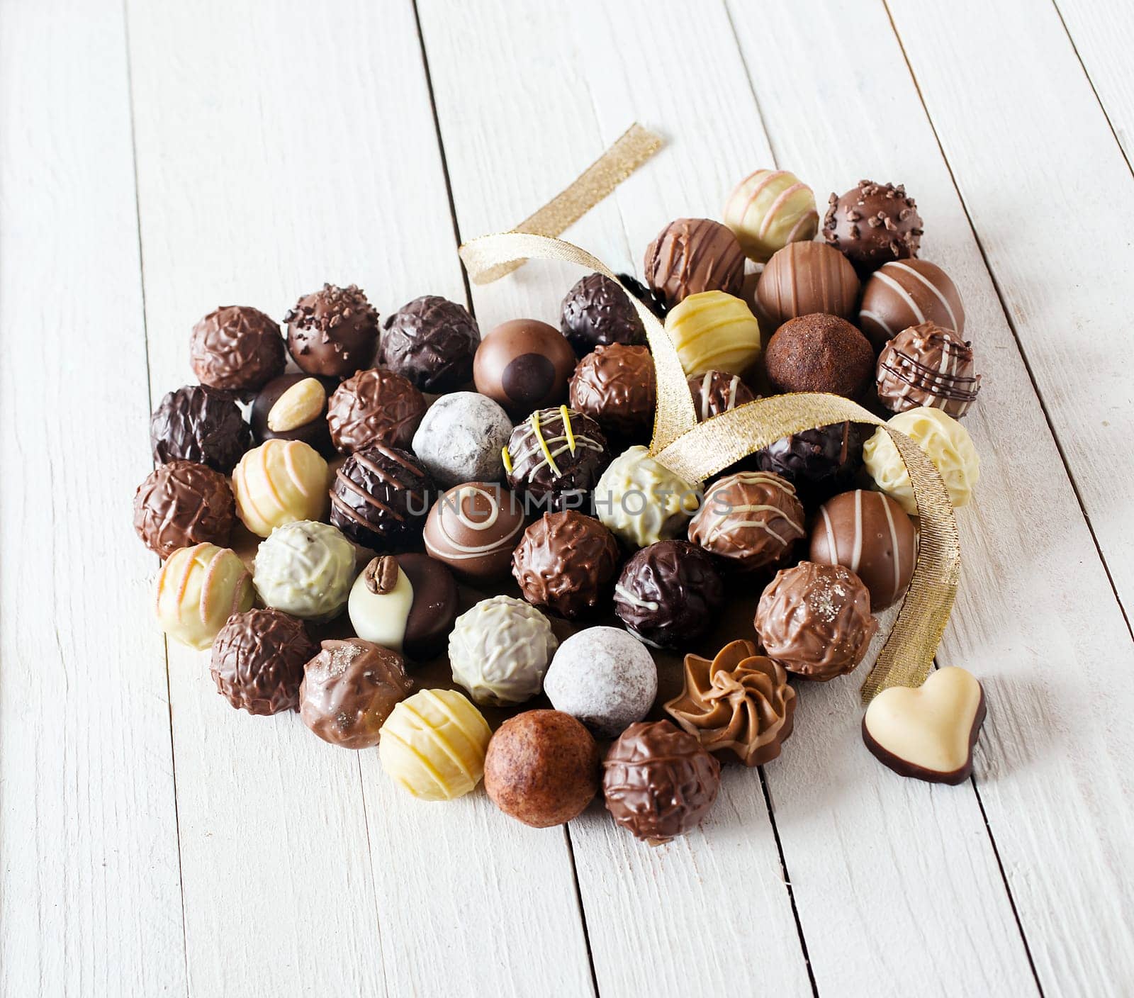 Close-up of a heart shape made with various types of chocolate truffles decorated with a curly golden ribbon over a white wooden table