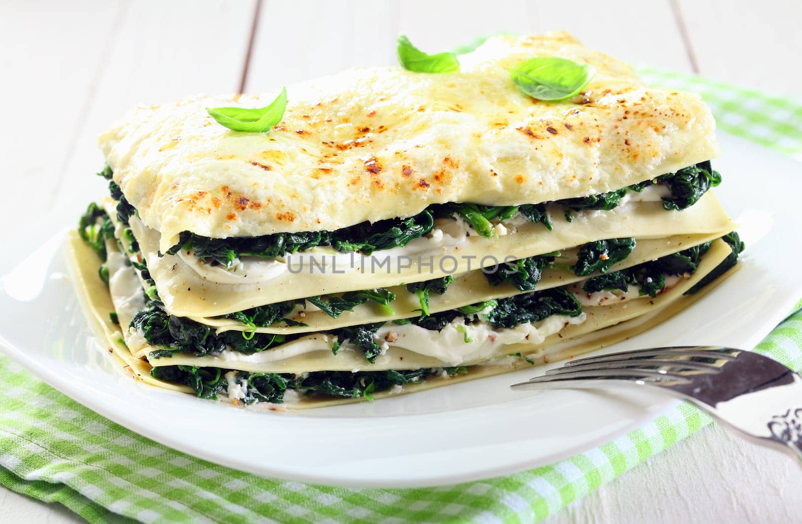 Close-up of a fresh baked vegetarian spinach lasagna on a plate, with a fork and over a napkin and an old white wooden table