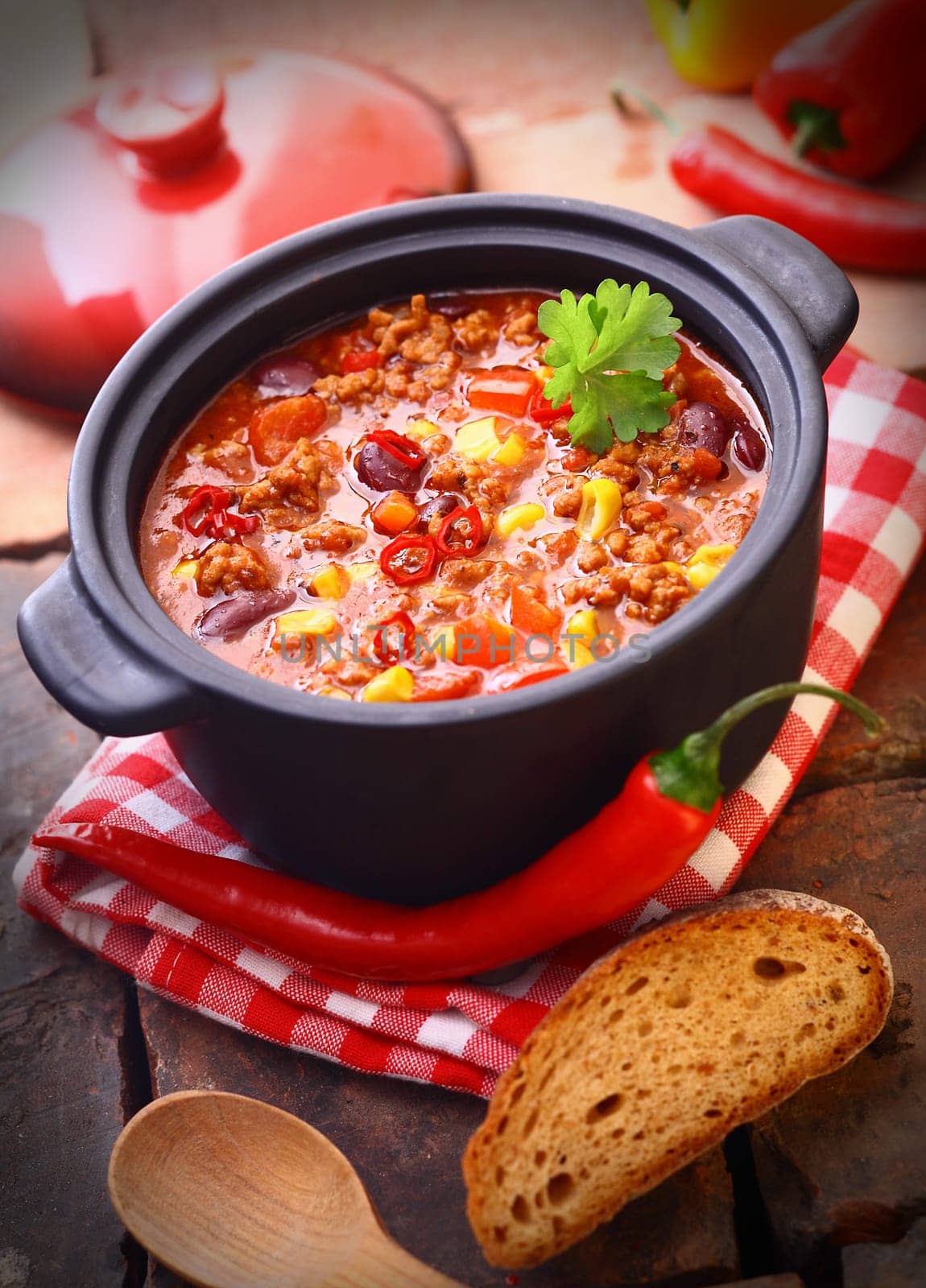 Hot and spicy fresh made Mexican chili still in an iron pot, with a raw chili-pepper and a slice of bread