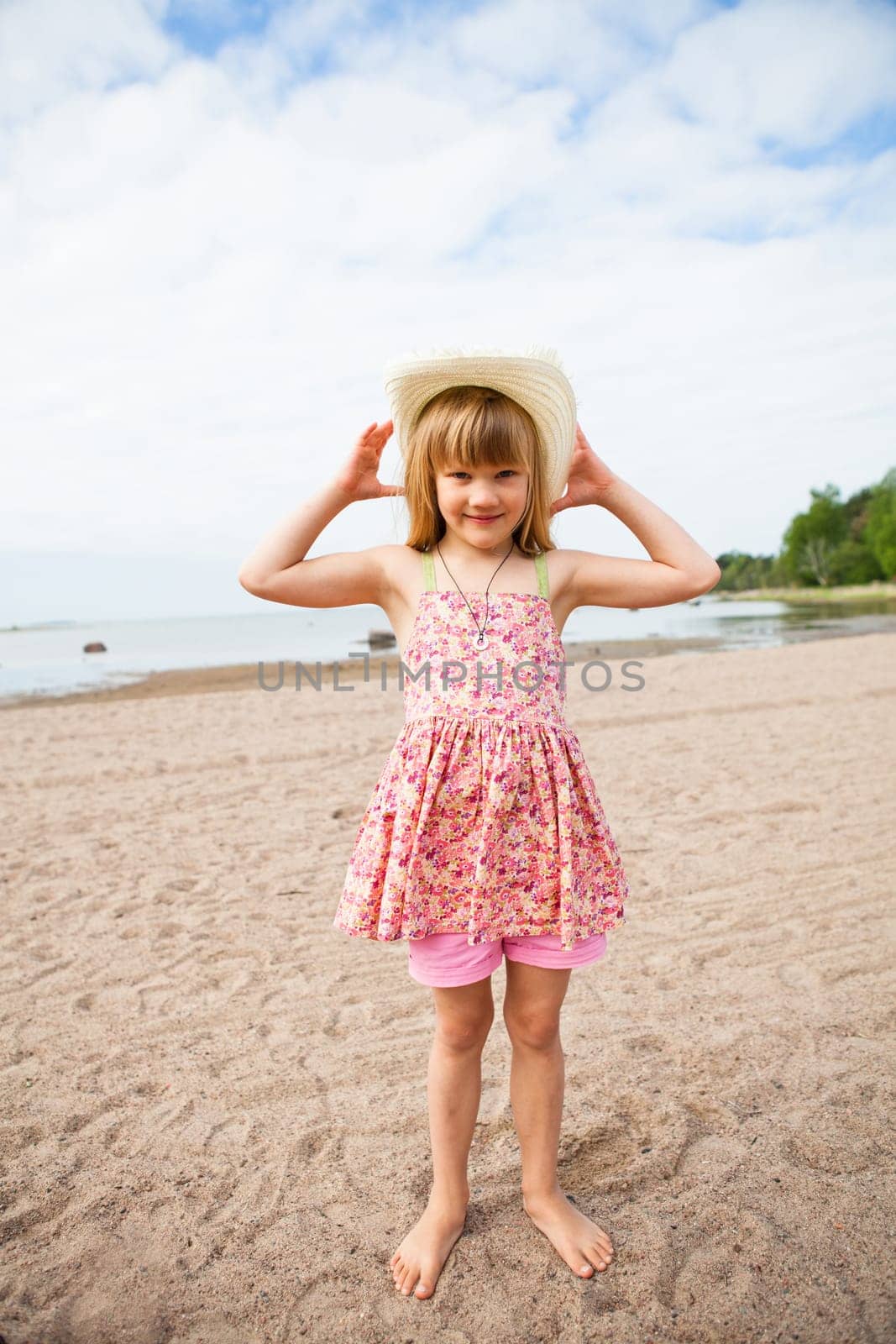 Smiling young barefoot girl wearing hat and pink patterned dress at beach