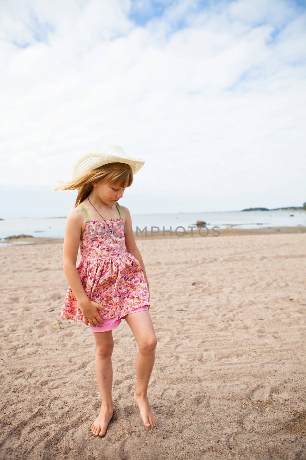 Thoughtful young barefoot girl wearing hat and pink patterned dress at beach with ocean in background