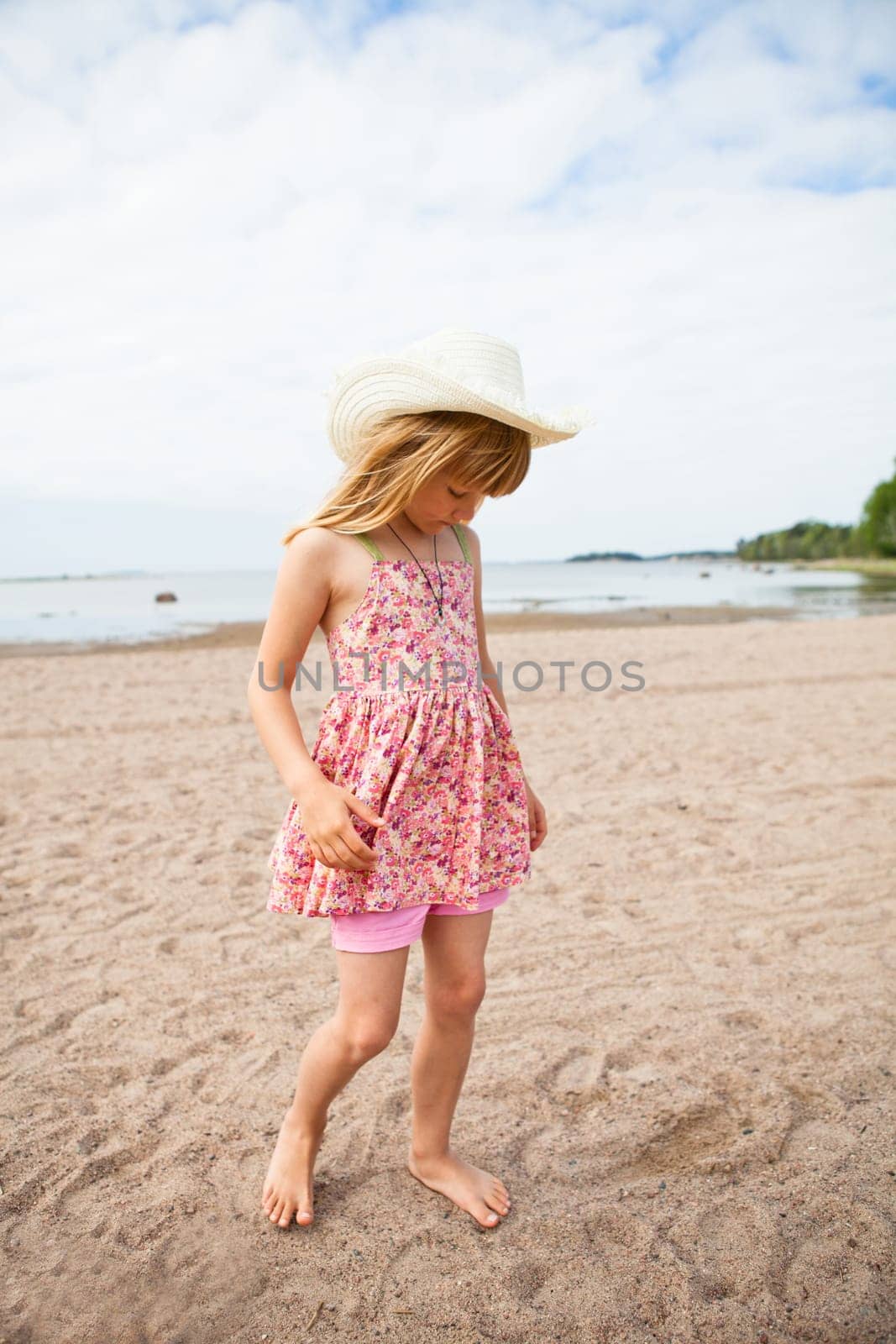 Thoughtful young barefoot girl wearing hat and pink patterned dress at beach with ocean in background