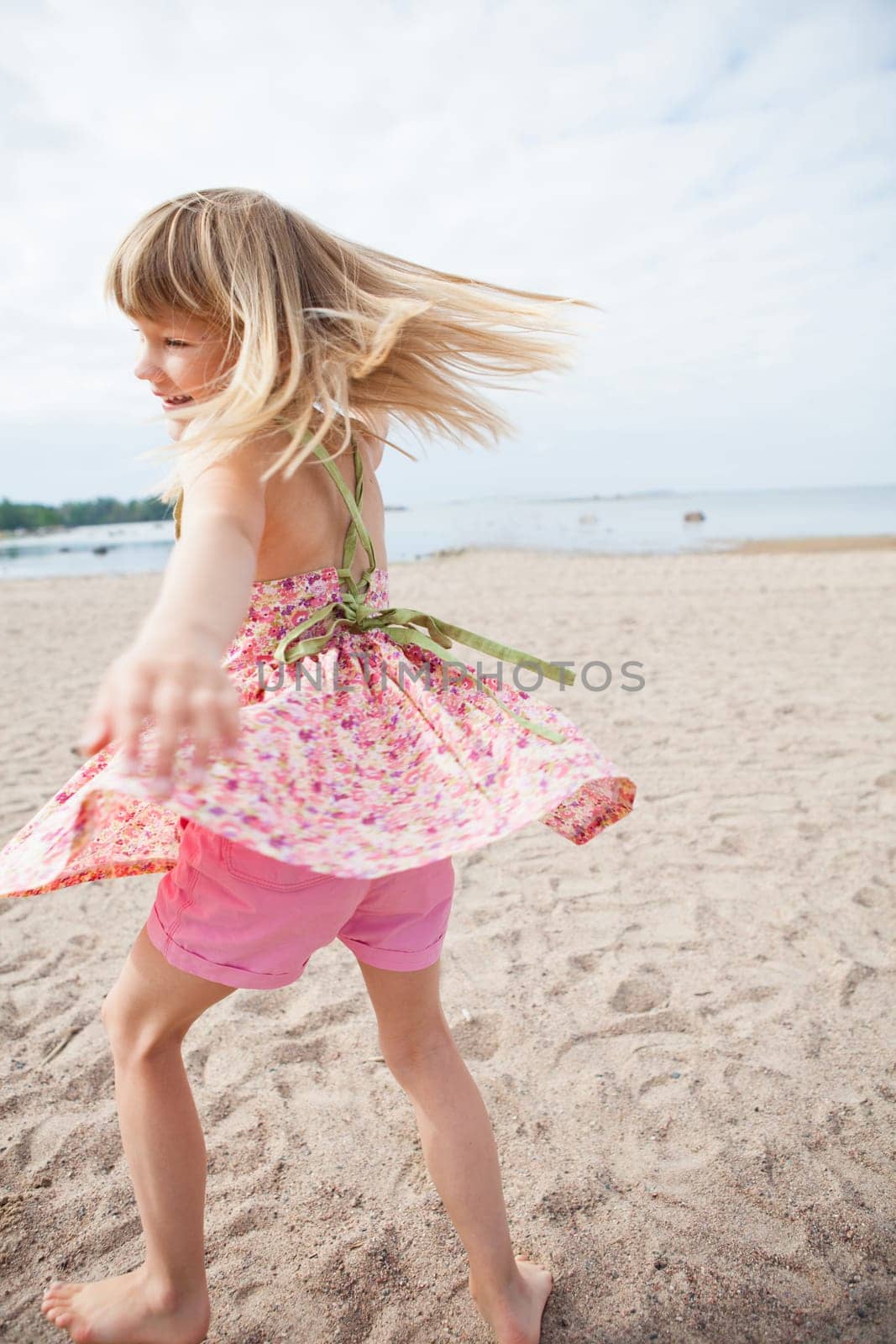 Smiling young barefoot girl with pink patterned dress and pants having fun at beach with ocean in background