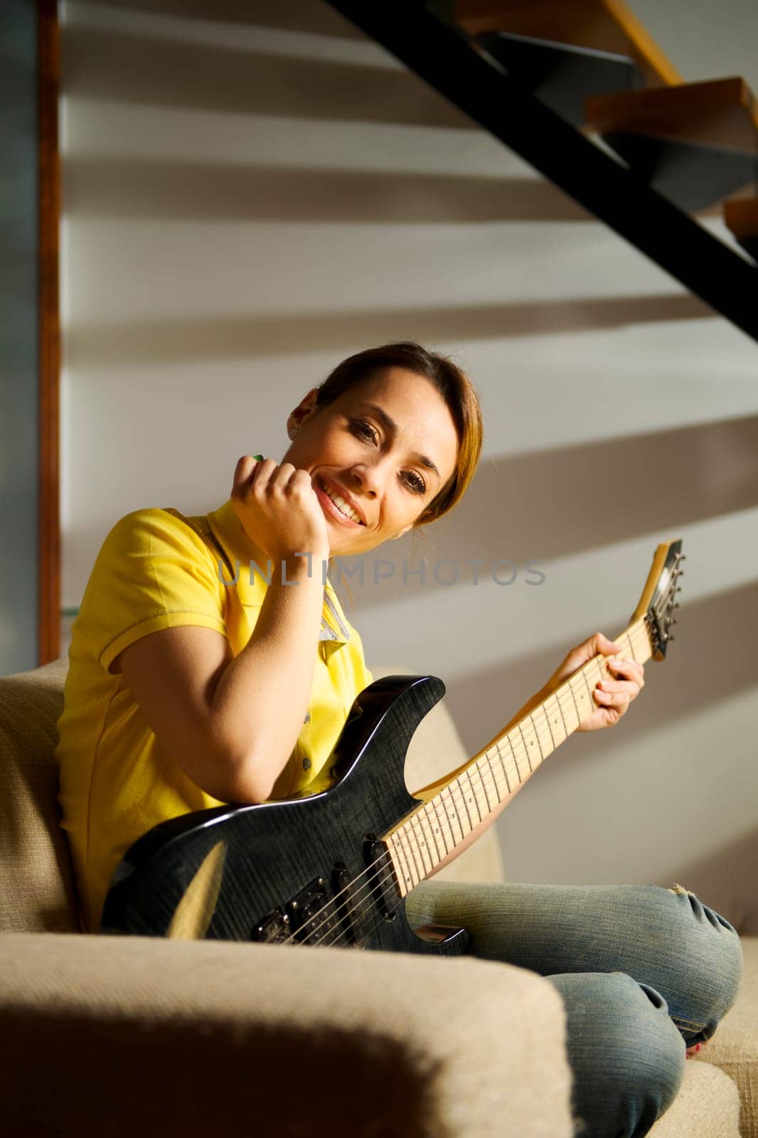 portrait of beautiful mid adult woman playing electric guitar at home, sitting on sofa