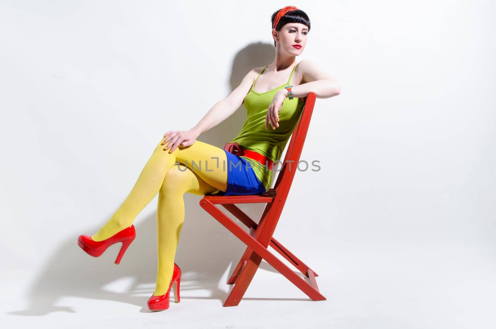 Young woman with black straight hair wears colorful mixed clothes and stands in front of a white background.