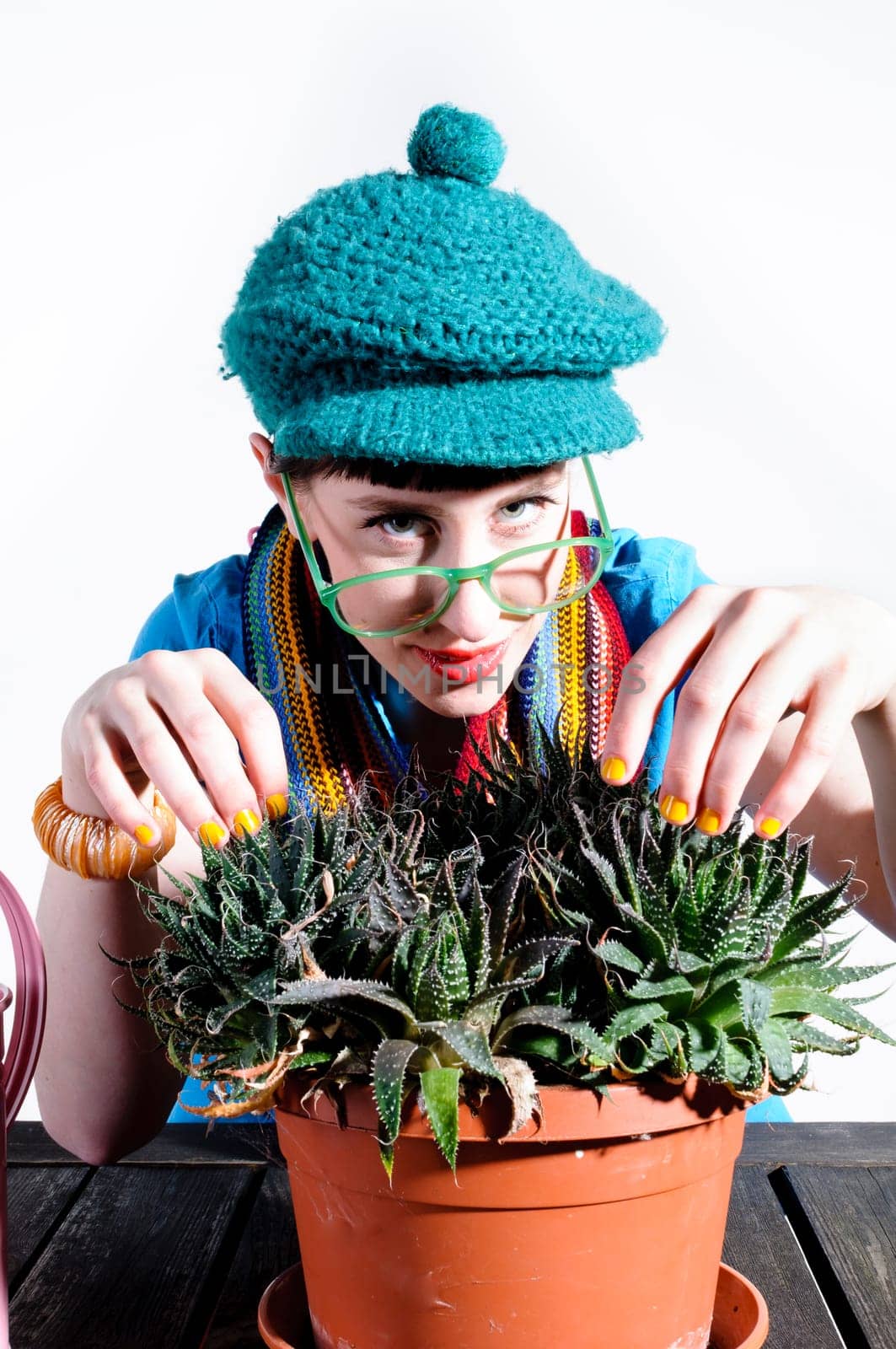 Young woman with black straight hair wears colorful mixed clothes and stands in front of a white background and devotedly pours a cactus.
