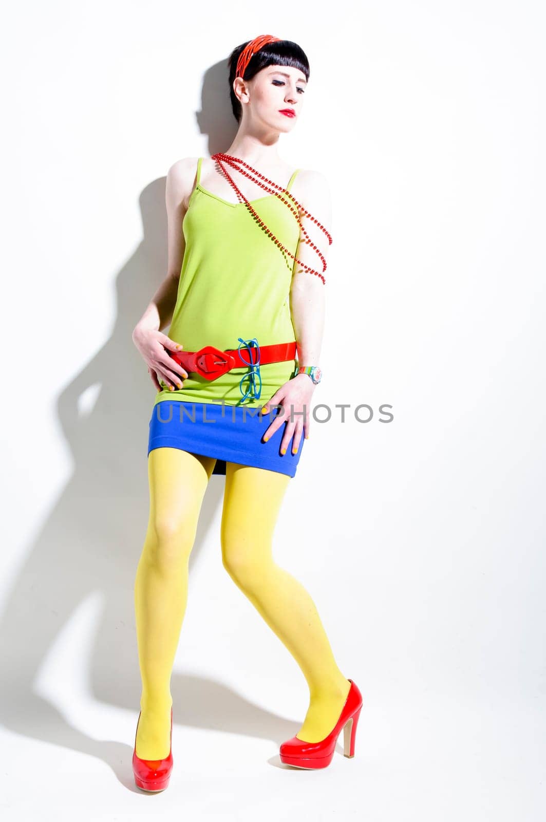 Young woman with black straight hair wears colorful mixed clothes and stands in front of a white background.