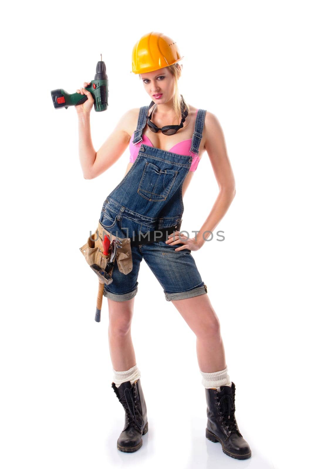 Young attractive woman wears denim dungarees,a tool belt and an orange construction helmet. She holds a cordless screwdriver and stands in front of a white background.