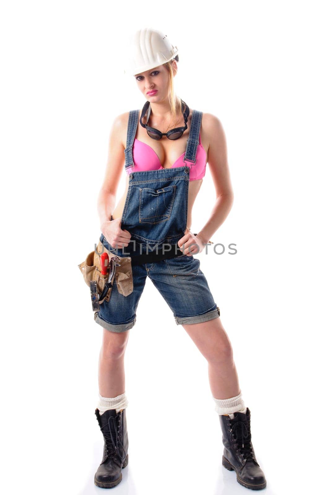 Young attractive woman wears denim dungarees,a tool belt and an orange construction helmet and stands in front of a white background.