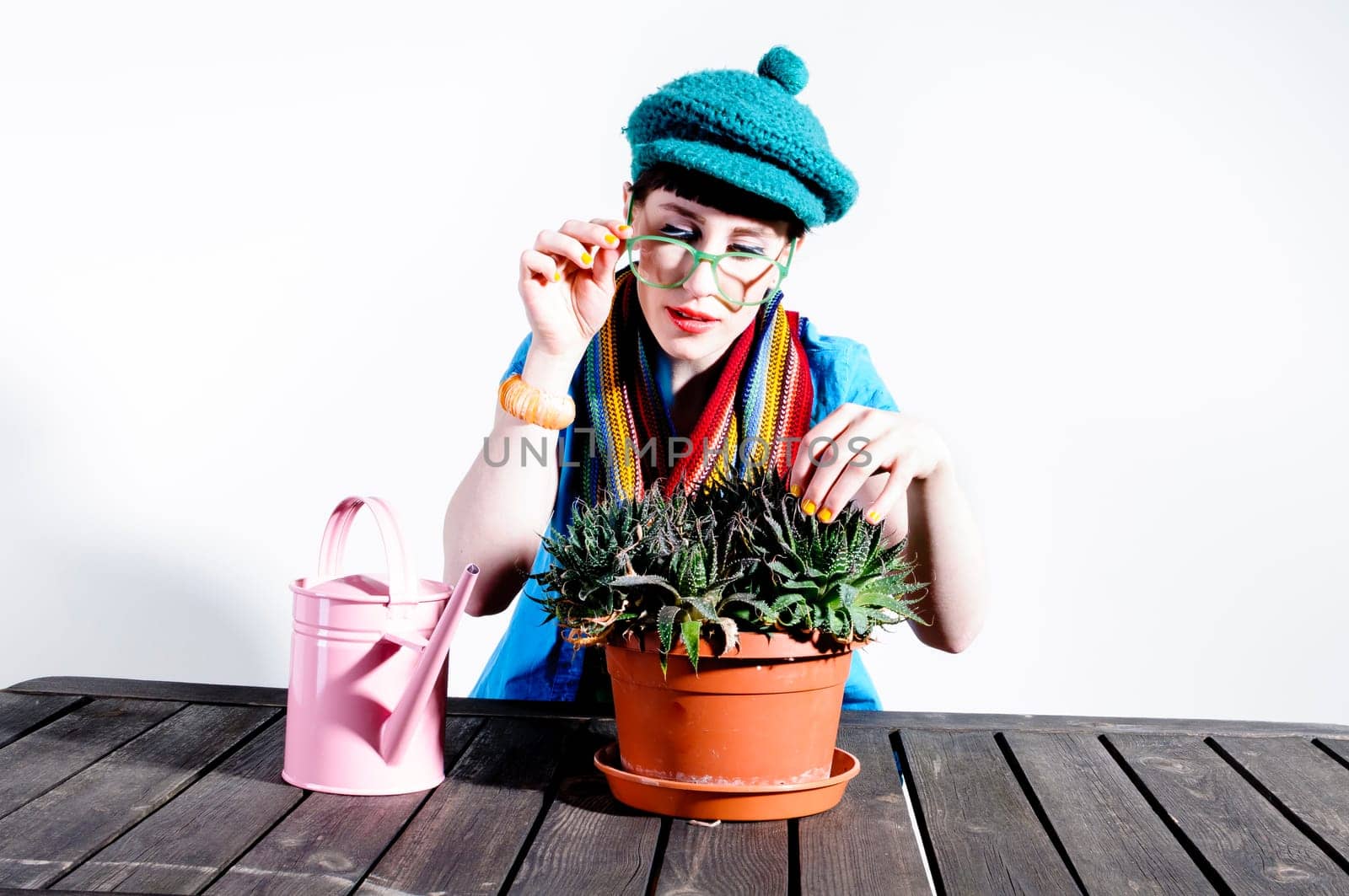 Young woman with black straight hair wears colorful mixed clothes and stands in front of a white background and devotedly pours a cactus.