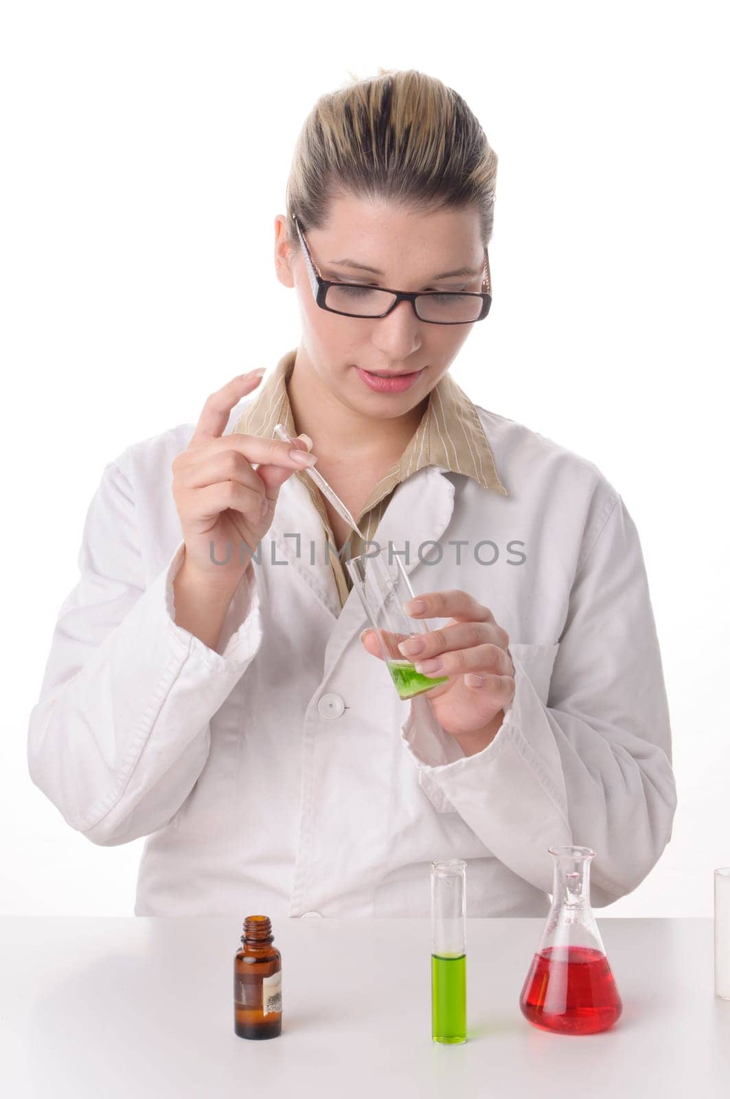 Attractive young chemical laboratory assistant with blond hair and glasses works with various colored liquids in test tubes,exposed against a white background. On the table are various colored chemicals.