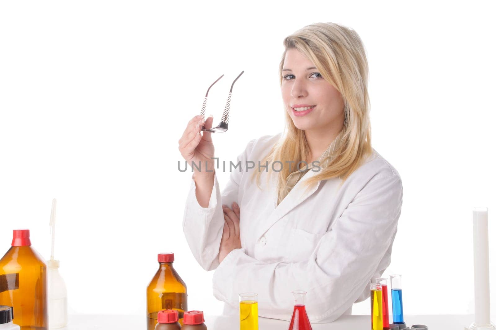 Attractive young chemists with blond hair and glasses wears a white coat. She stands contentedly and proudly with folded arms behind a table with various chemicals,exposed against a white background
