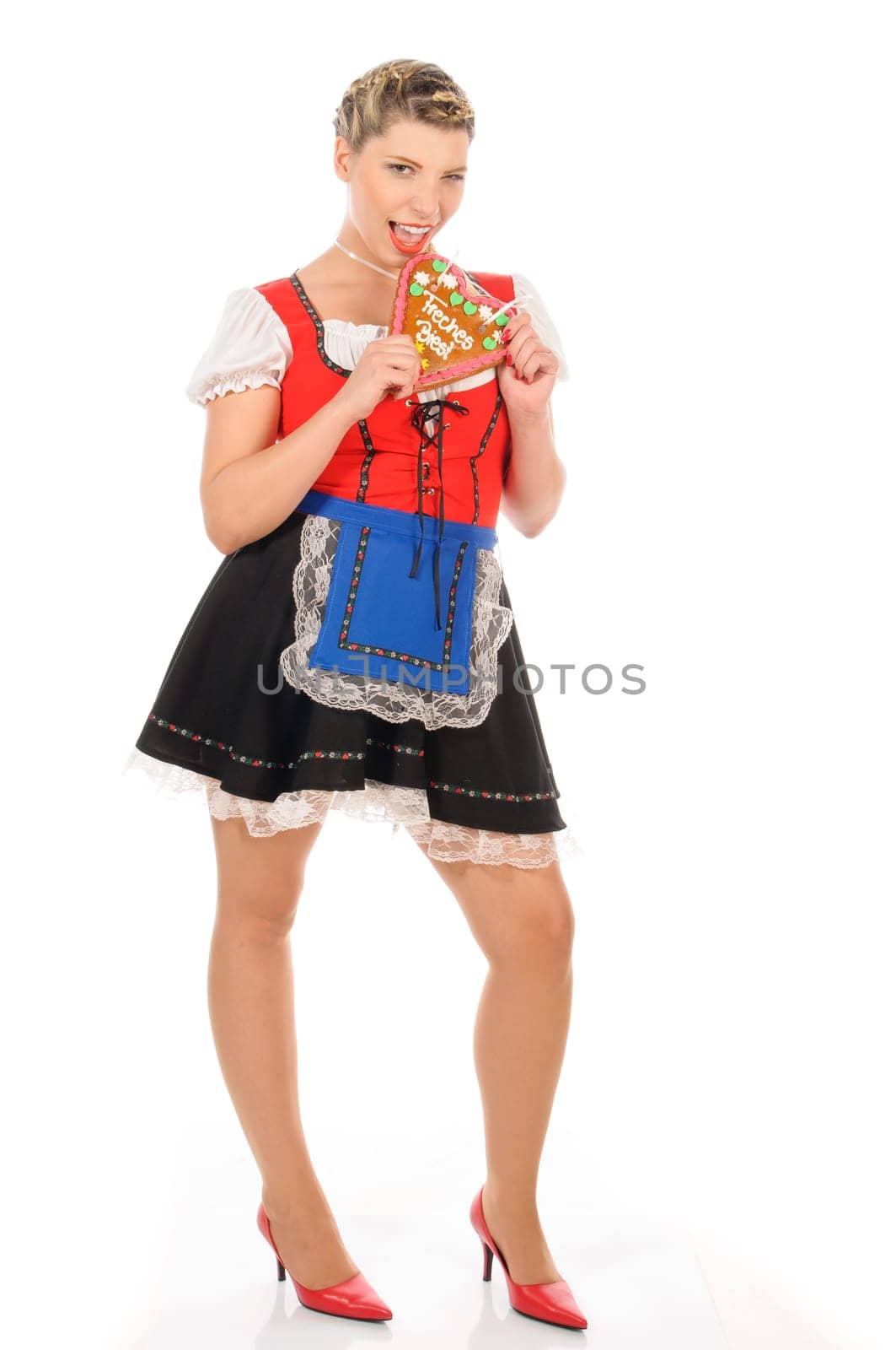 Young attractive woman with blond hair and pigtails wears a colorful dirdl dress and holds a gingerbread heart with the inscription 'Naughty Beast' in her hand,exposed against a white background.