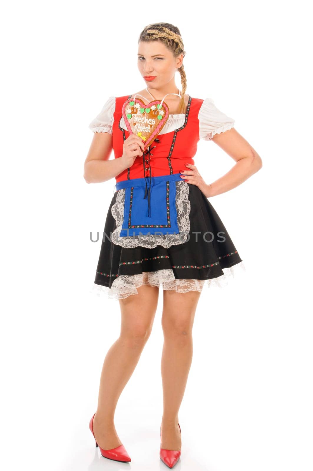 Young attractive woman with blond hair and pigtails wears a colorful dirdl dress and holds a gingerbread heart with the inscription 'Naughty Beast' in her hand,exposed against a white background.