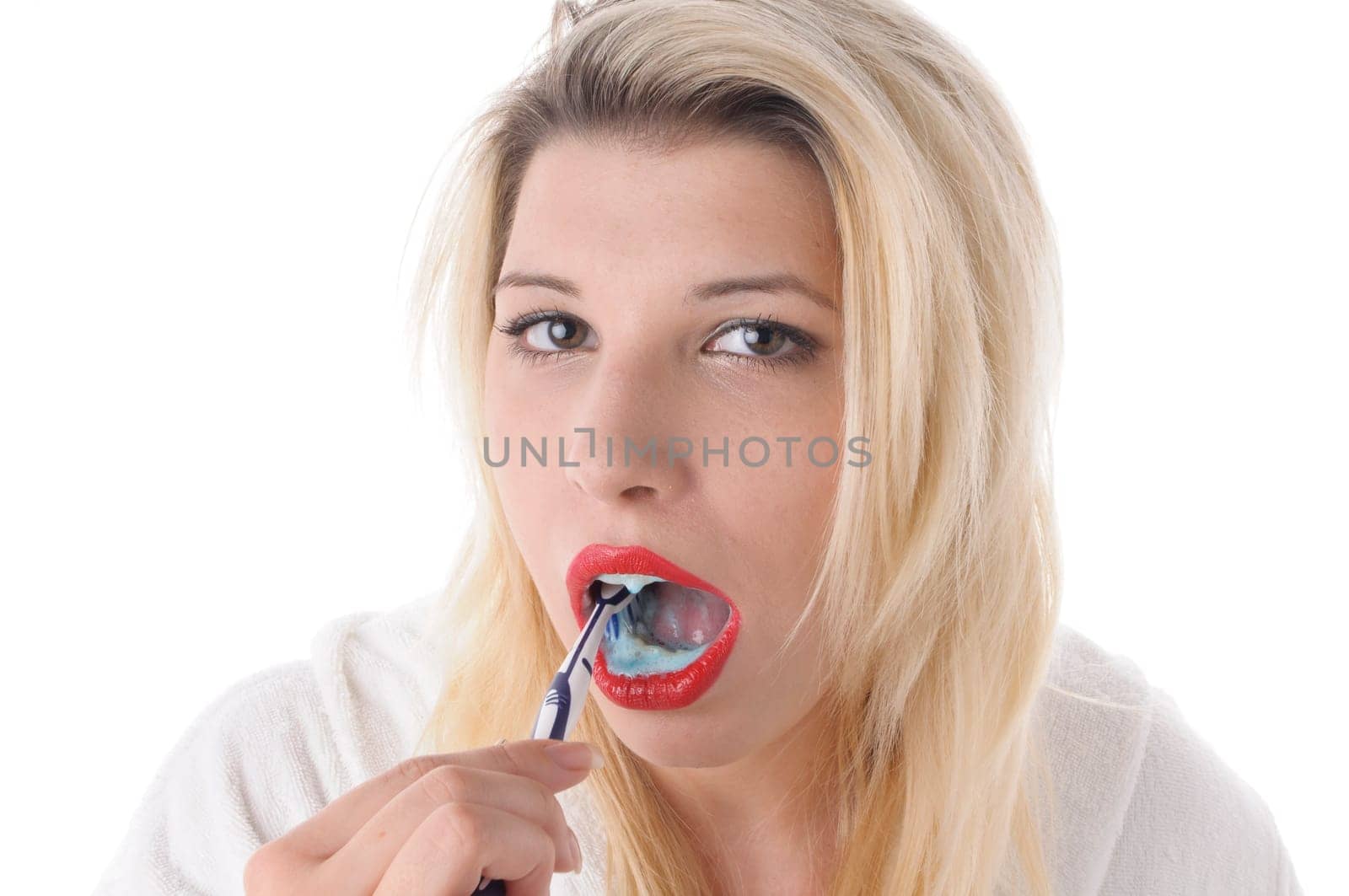 Attractive young woman with long blonde hair wears a white bathrobe and brushes her teeth with a toothbrush,exposed against a white background.