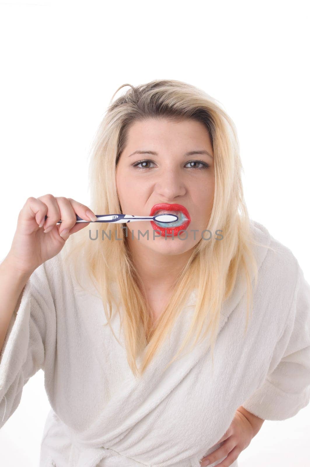 Attractive young woman with long blonde hair wears a white bathrobe and brushes her teeth with a toothbrush,exposed against a white background.