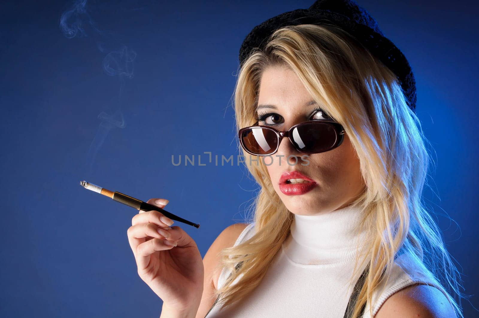 Attractive young woman with long blonde hair wears a tight sleeveless turtleneck top,black shorts with suspenders and high heels. She casually smokes a cigarette with a cigarette tip,against a dark background.