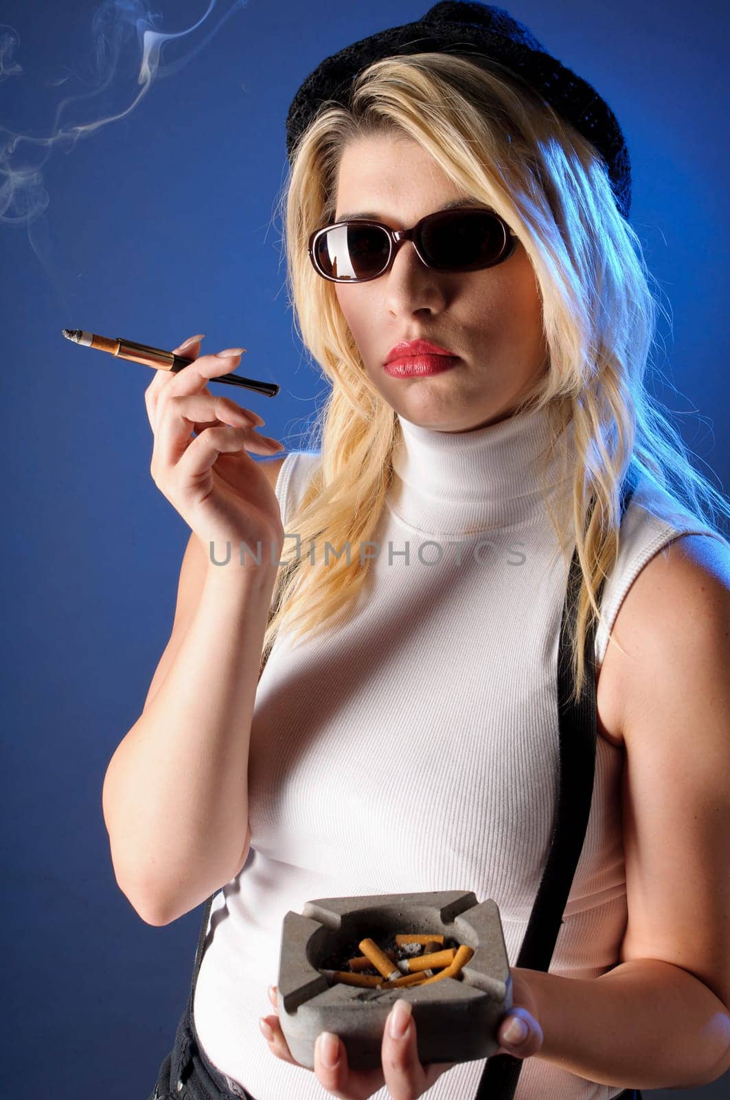 Attractive young woman with long blonde hair wears a tight sleeveless turtleneck top,black shorts with suspenders and high heels. She casually smokes a cigarette with a cigarette tip,against a dark background.
