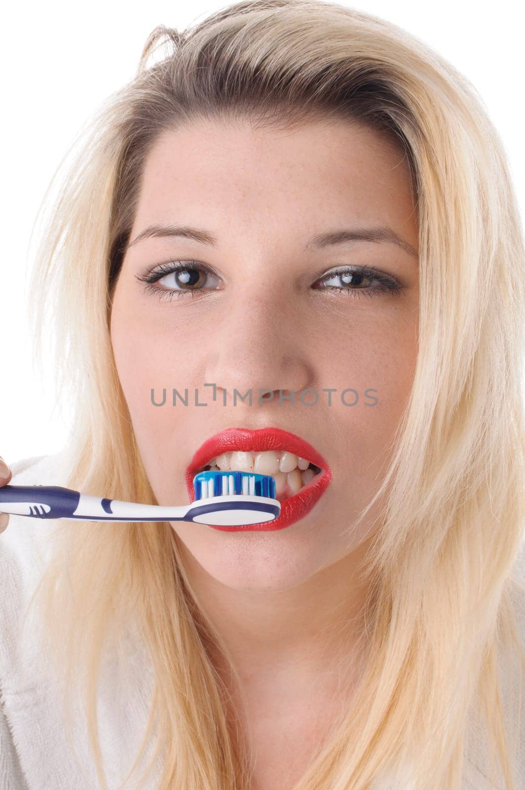 Attractive young woman with long blonde hair wears a white bathrobe and brushes her teeth with a toothbrush,exposed against a white background.