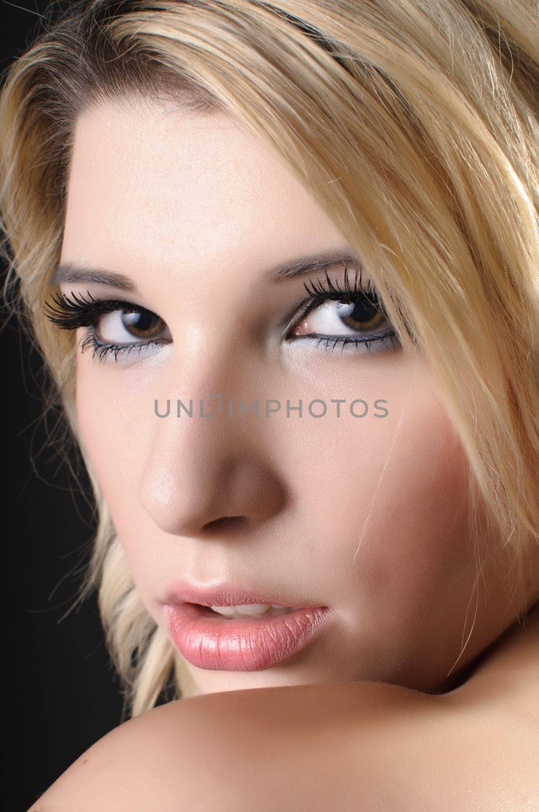 A pretty young woman with blond hair sits in front of a dark background. She is covered only with a white cloth and shows her beautiful,naked back to the camera.
