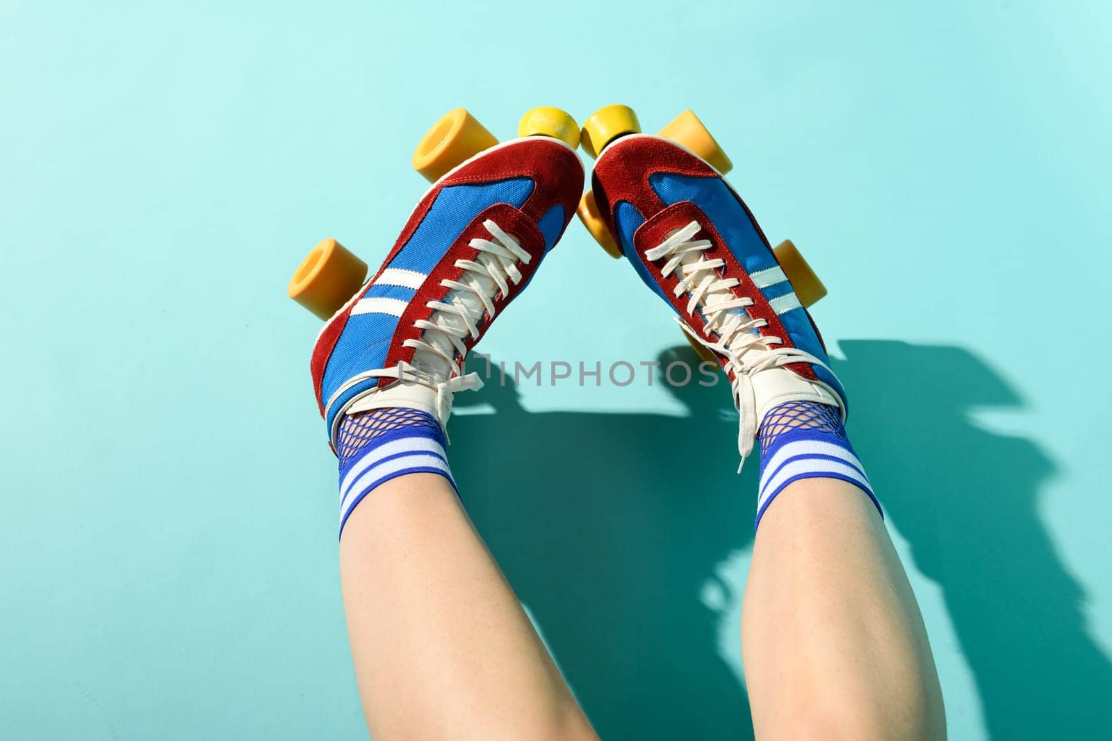 First person POV of the legs of a woman wearing colorful red and blue roller skates with yellow wheels and anklet socks over a blue studio background with shadow