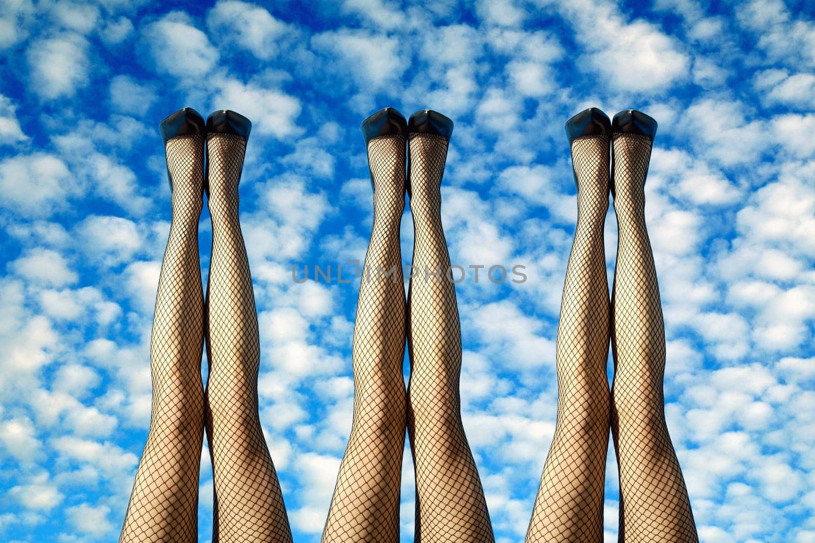 3 pairs of sexy female legs in black fishnet stockings and stilettos raised up side by side against a cloudy sunny blue sky in a conceptual image