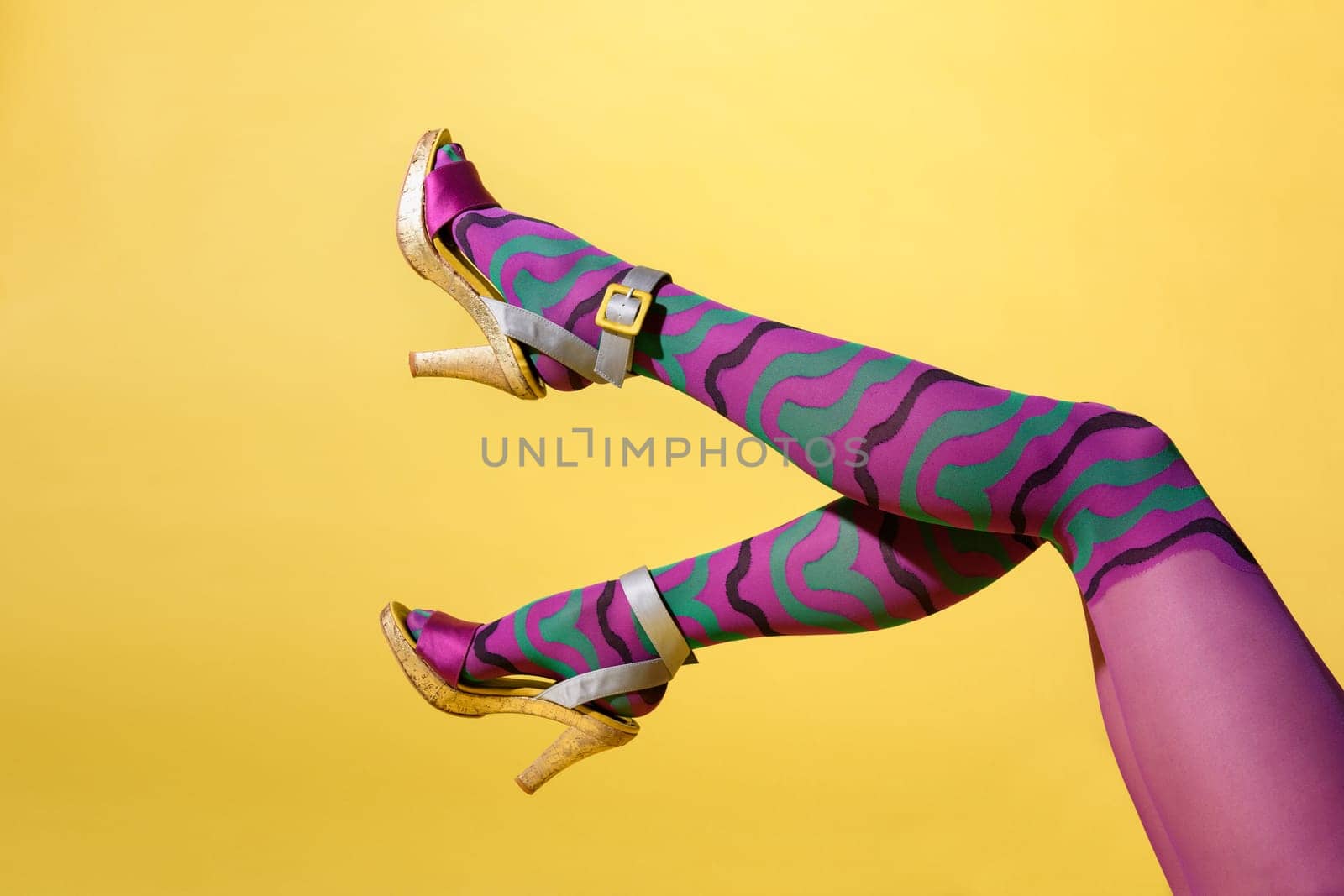 Waving sexy female legs in multicolored purple collant and high heeled shoes with buckle in a side view over a yellow background with copyspace