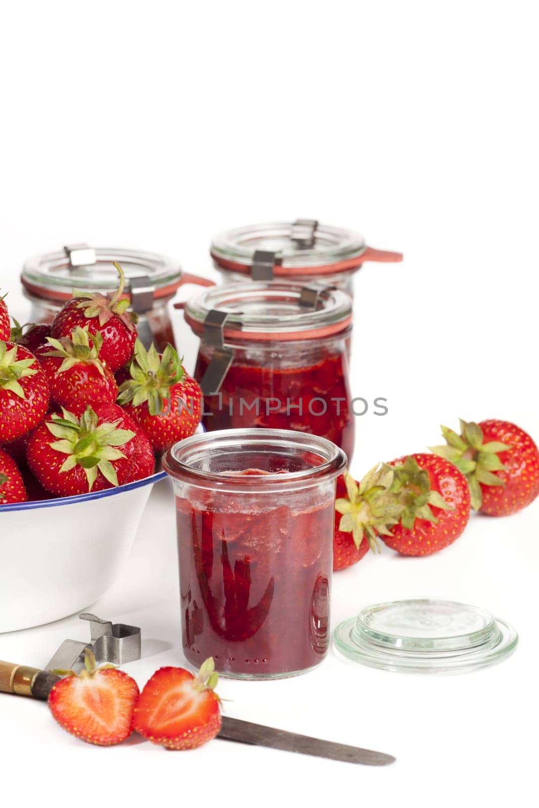 Freshly harvested strawberries in a white enamel bowl and homemade jam in jars against a white background