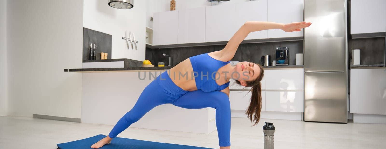 Image of fit young woman doing workout, stretching her body, doing fitness, aerobics workout on yoga mat, standing in asana in living room, training at home.