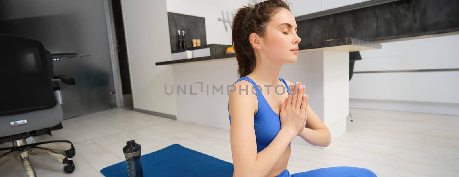 Sport and recreation. Close up portrait of beautiful girl does yoga at home, clasps her hands and meditates, sits on rubber mat in kitchen, makes asana, relaxing.
