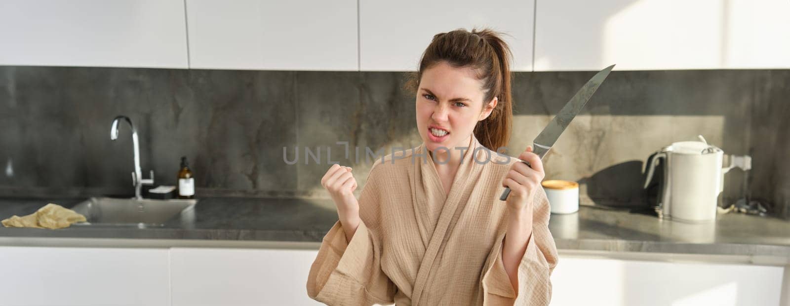 Portrait of annoyed woman with knife, angry while cooking in the kitchen, frustrated while doing house chores and preparing food for family, standing in bathrobe.