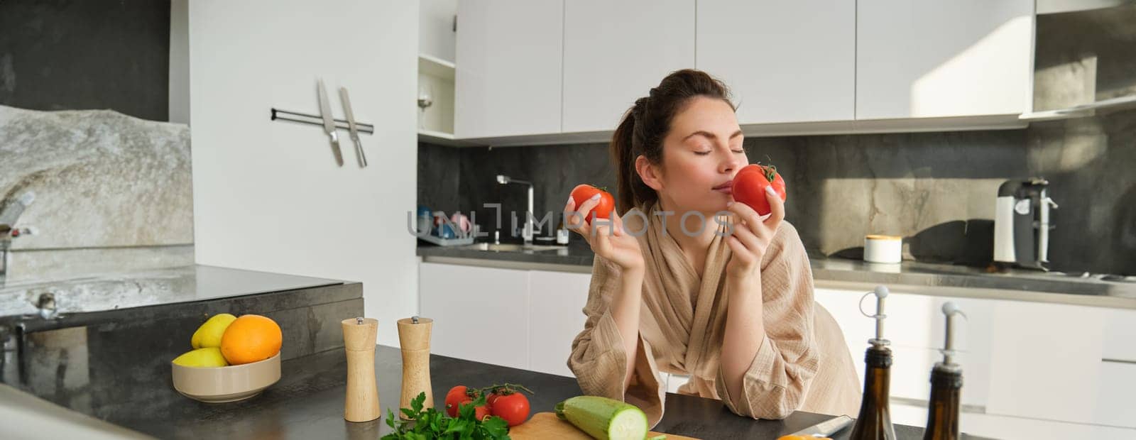 Portrait of beautiful young woman smelling tomatoes, cooking salad from fresh vegetables bought in market, preparing food for family, lead active and healthy lifestyle, standing in kitchen.