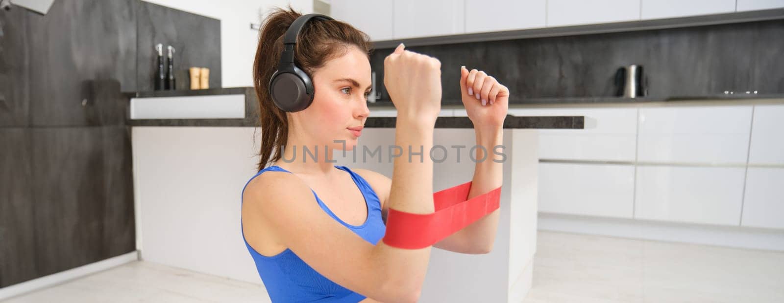 Portrait of determined beautiful woman, listening music, workout with elastic resistance band, doing exercises on arms, wearing sportsbra.