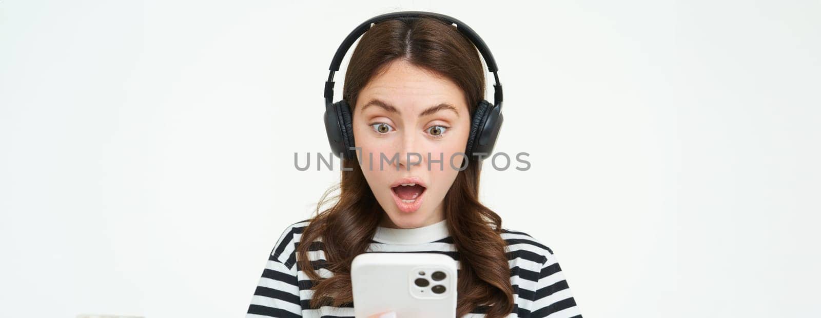 Portrait of young woman in wireless headphones, looks shocked at smartphone screen, reads message on mobile phone with dropped jaw, gasping from surprise, white background by Benzoix