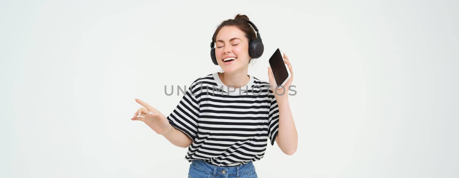 Girl with smartphone dancing, listening to music in wireless headphones, using mobile phone streaming app to enjoy favourite songs, isolated over white background by Benzoix