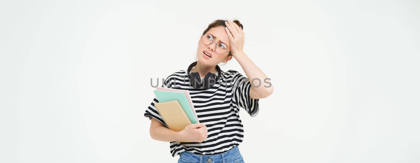 Portrait of compllicated, sad young woman in glasses, carries homework notebooks, facepalms, looks disappointed, stands over white background by Benzoix