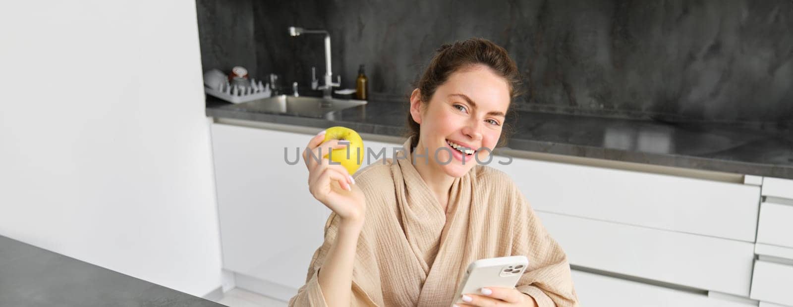 Portrait of happy, beautiful woman smiling, eating an apple in the kitchen, sitting at home in bathrobe, holding smartphone, using mobile phone by Benzoix