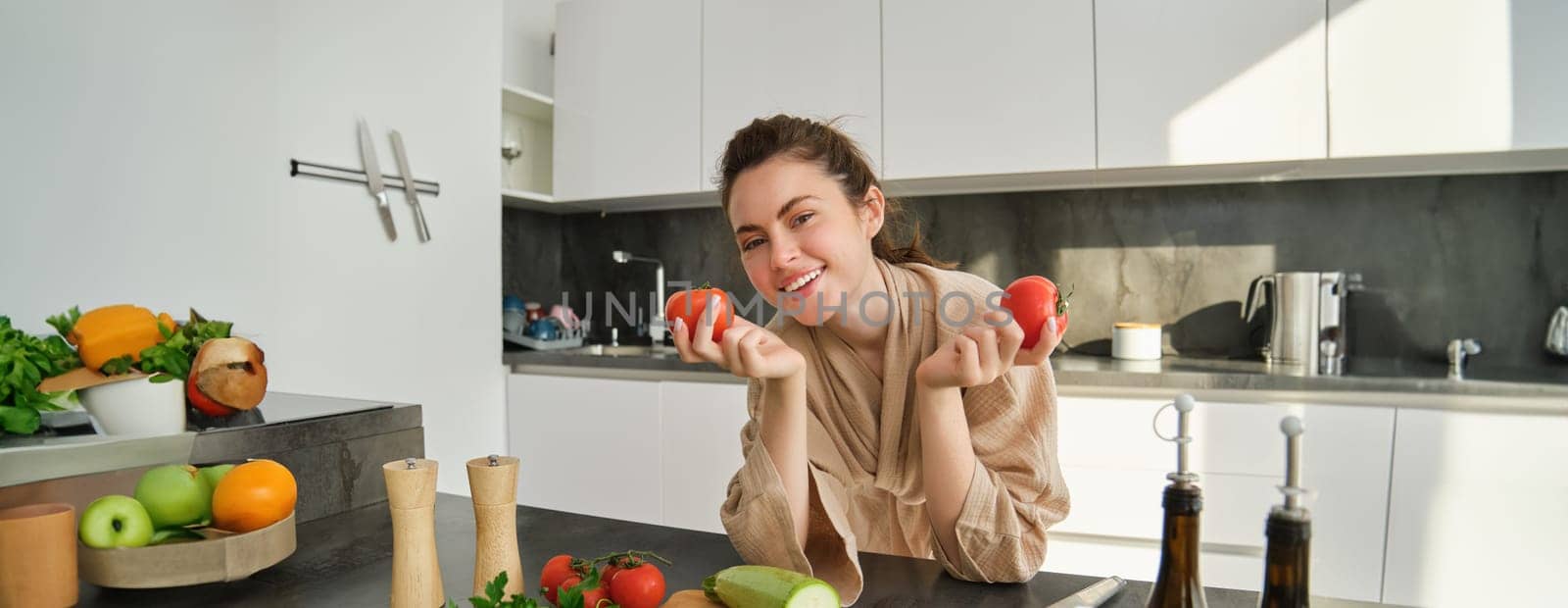 Portrait of beautiful woman holding tomatoes, standing in the kitchen in bathrobe and cooking vegetarian meal, laughing while preparing food from fresh vegetables by Benzoix