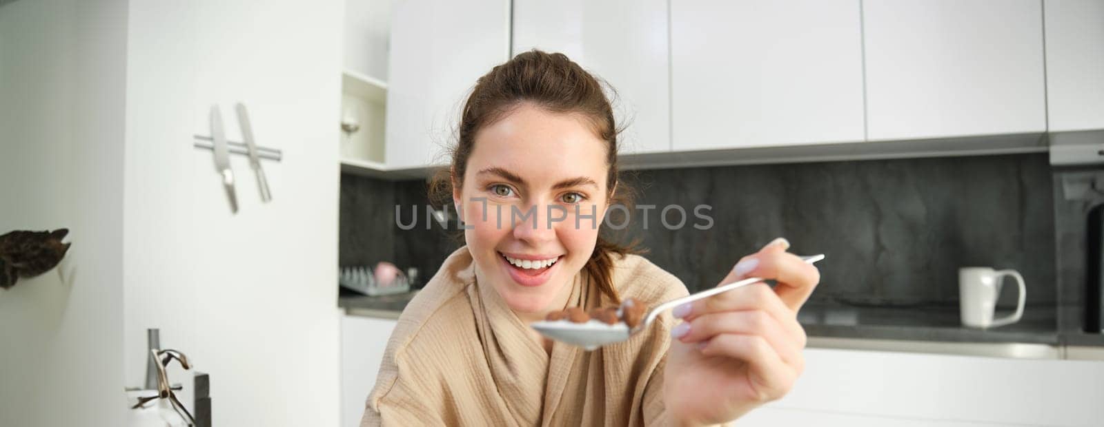 Portrait of happy young woman leans on kitchen worktop and eating cereals, has milk and bowl in front of her, having her breakfast, wearing bathrobe.