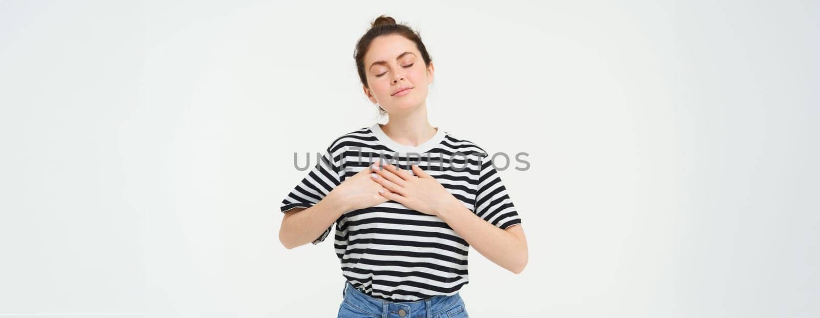 Passionate girl holds hands on her heart and smiles, stands over white background.