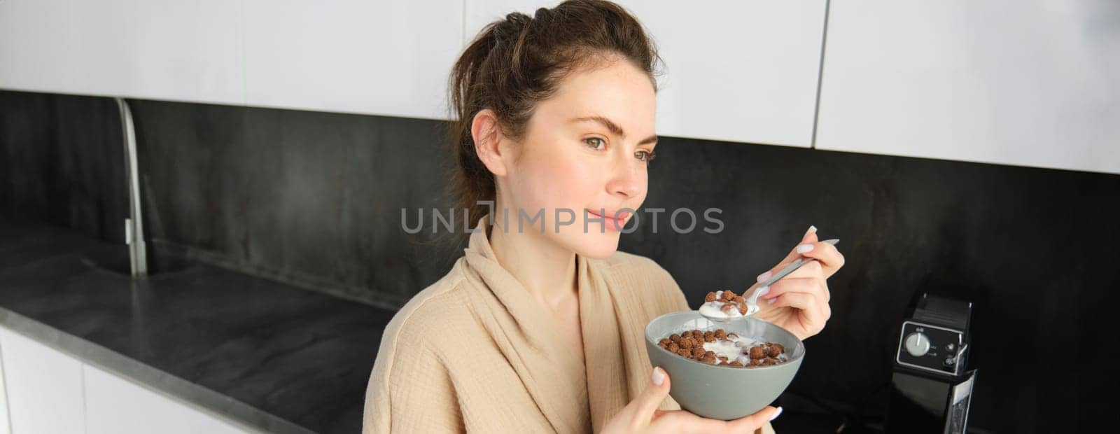 Good-looking brunette woman eating her breakfast, standing in kitchen near worktop and holding bowl of cereals with milk, enjoying her morning, wearing cosy bathrobe.