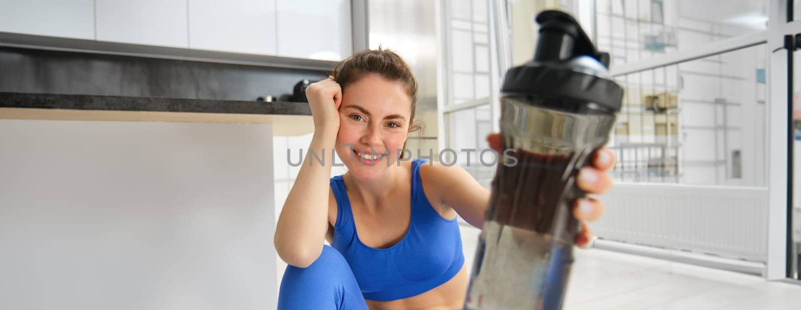 Young positive girl does workout from home, gives you water bottle to drink during training exercises, sits on yoga mat floor in living room.