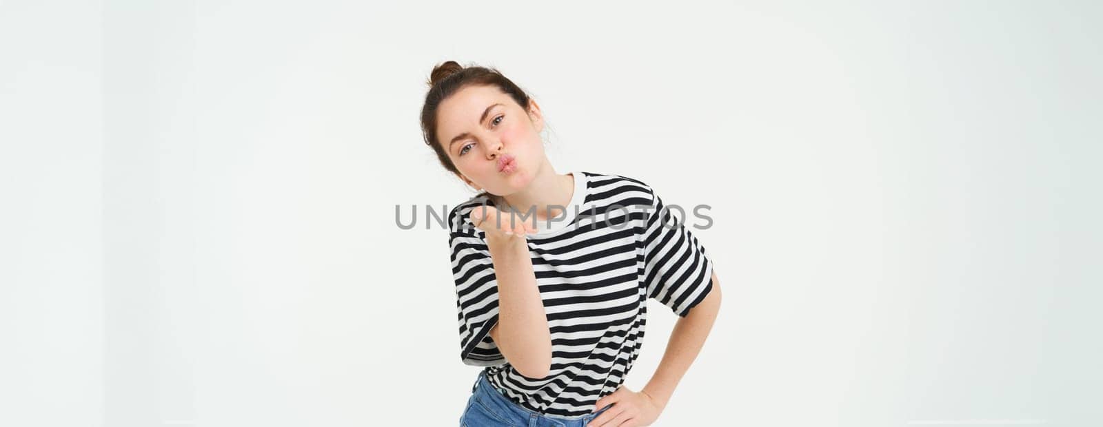 Portrait of beautiful brunette woman sends air kiss at camera, holds palm near lips, isolated against white background by Benzoix