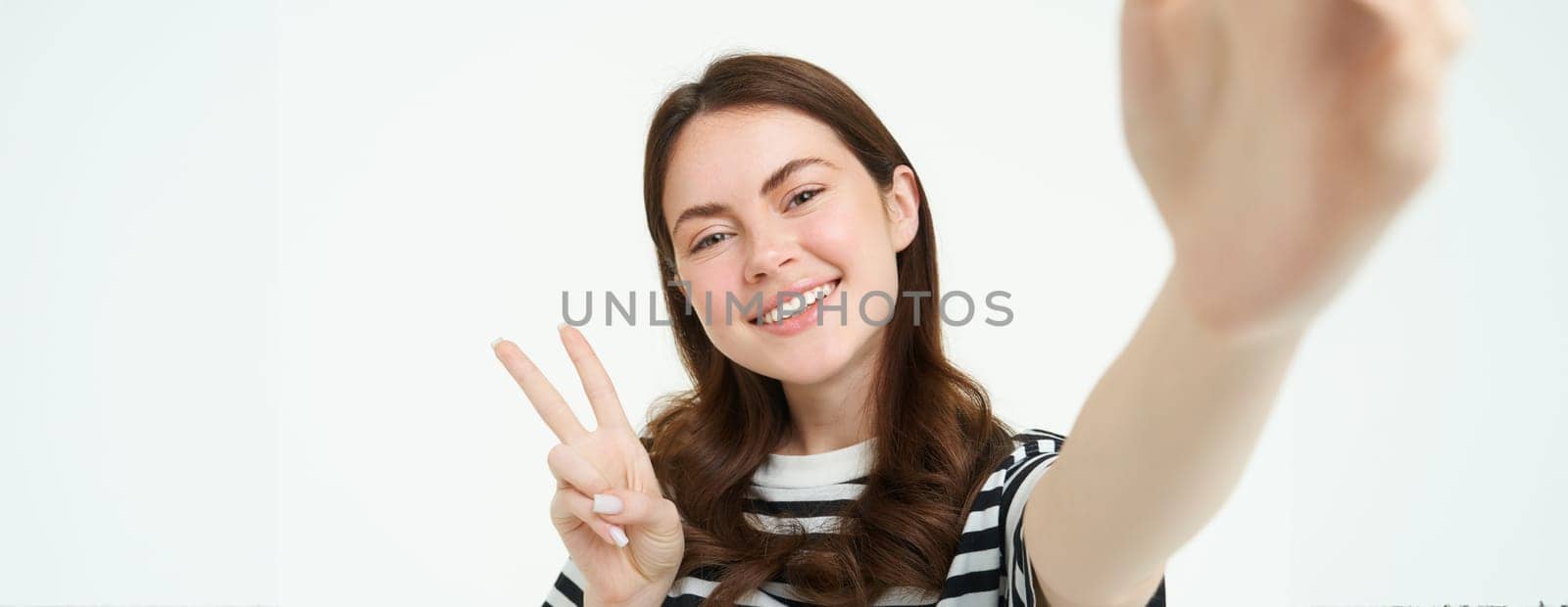 Smartphone view, woman holding mobile phone camera and taking selfie with peace, v-sign gesture, smiling at camera, posing over white background by Benzoix