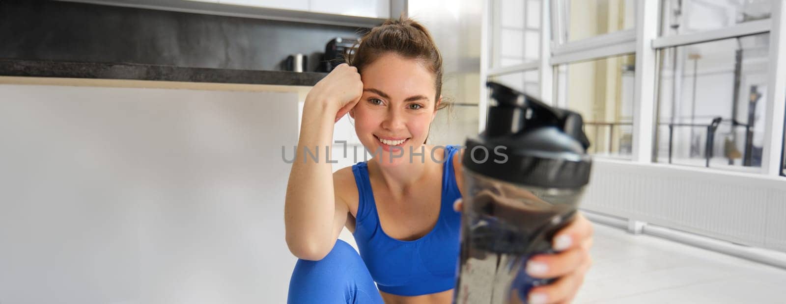 Smiling fitness girl drinks water in the middle of training session, smiling and looking happy, workout from home in blue sportsbra by Benzoix