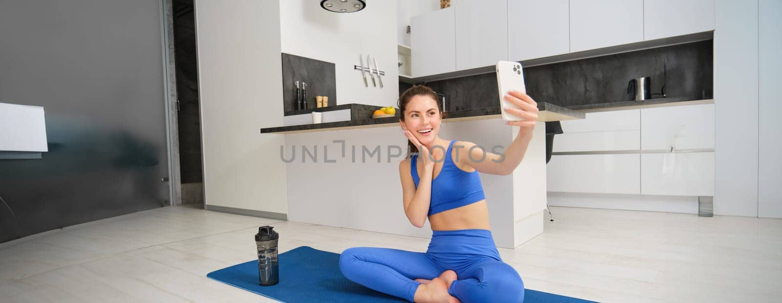 Young smiling woman, fitess girl doing workout from home, sits on yoga mat with smartphone, taking selfie, records sport vlog indoors.