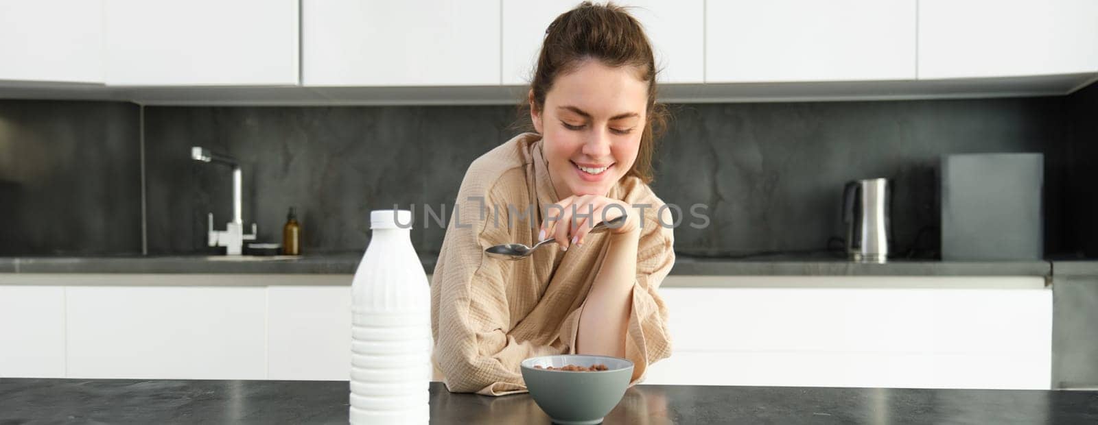 Portrait of beautiful young and healthy woman in bathrobe eats her breakfast in kitchen, has cereals with milk and smiling.