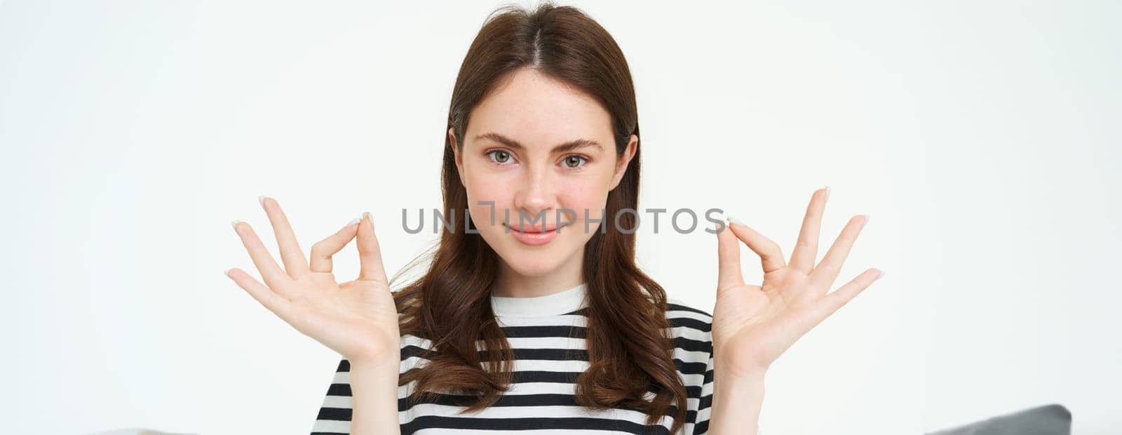 Portrait of woman smiling with confidence, showing okay, zero sign, meditating, standing over white background by Benzoix